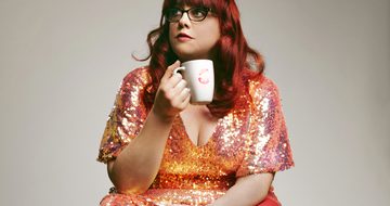 A lady with long red hair wearing a pink and orang sparkly dress, holding a mug, sitting against a grey background