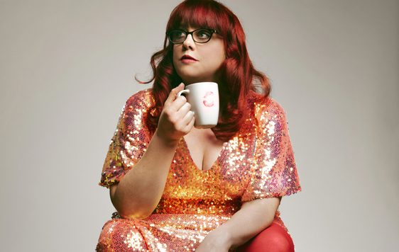 A lady with long red hair wearing a pink and orang sparkly dress, holding a mug, sitting against a grey background