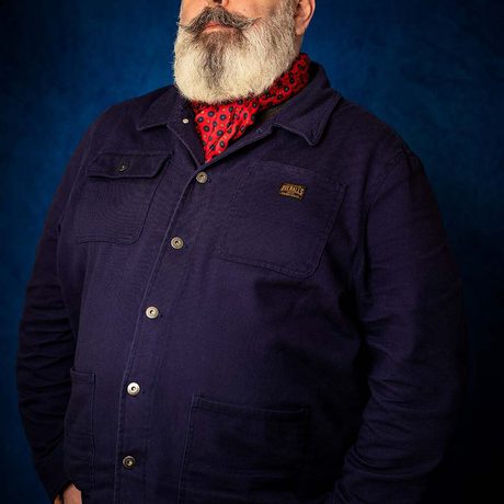 Picture of a man with a grey beard and moustache wearing a blue shirt with red scarf against a dark blue background