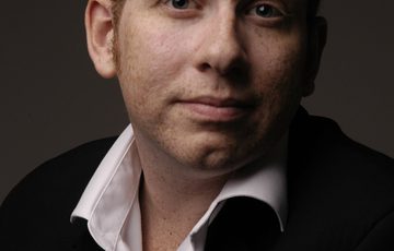 Portait of a man wearing a dark jacket and white shirt against a plain dark brown background