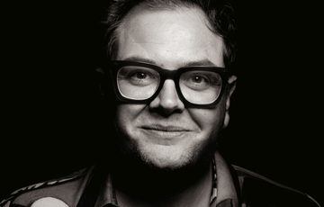 A black and white portrait of a man wearing glasses against a black background