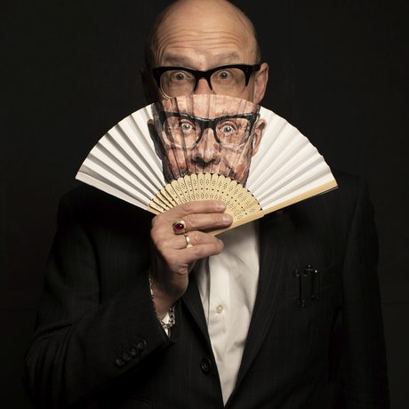 A man with glasses wearing a suit holding a fan with his face printed on it, against a black background