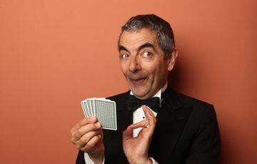 A man in a dinner suit holding a pack of playing cards sat at a green table with a terracotta background