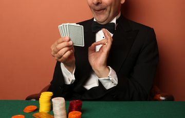 A man in a dinner suit holding a pack of playing cards sat at a green table with a terracotta background