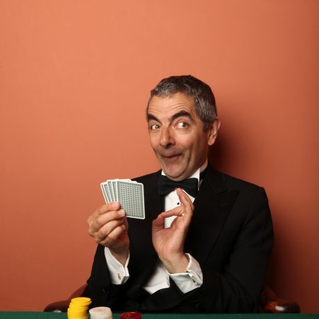 A man in a dinner suit holding a pack of playing cards sat at a green table with a terracotta background