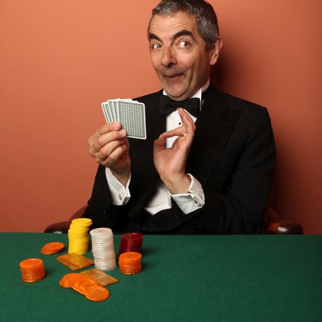 A man in a dinner suit holding a pack of playing cards sat at a green table with a terracotta background