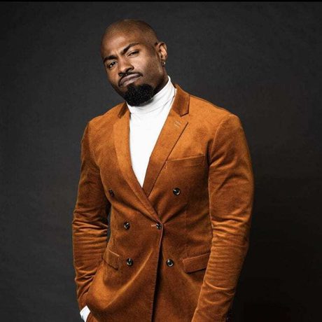 Darren Harriott wearing a brown/orange suit against a black background.