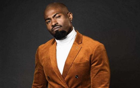 Darren Harriott wearing a brown/orange suit against a black background.