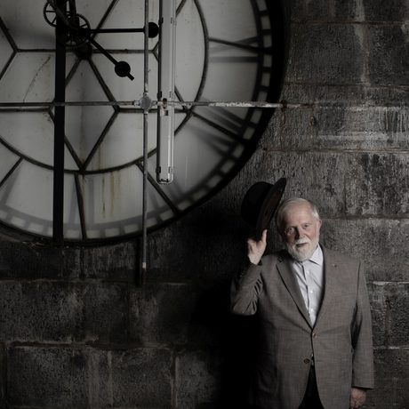a man holding a bowler hat standing in front of a stone wall with a large clock to the side