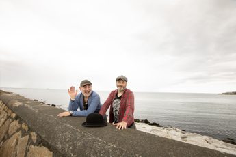 Two men wearing caps and a blue and other with red top, standing behind a sea wall with a black bowler hat in front of them.