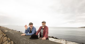 Two men wearing caps and a blue and other with red top, standing behind a sea wall with a black bowler hat in front of them.