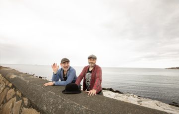Two men wearing caps and a blue and other with red top, standing behind a sea wall with a black bowler hat in front of them.