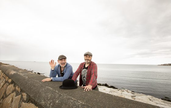 Two men wearing caps and a blue and other with red top, standing behind a sea wall with a black bowler hat in front of them.