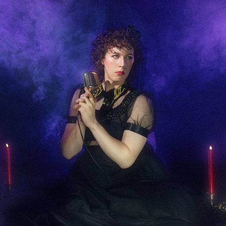 Sam Nicorest holding a microphone with candelabra's either side and a smokey purple background.