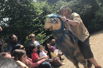 A person holding a cream and blue dinosaur puppet infront of a audience of people.