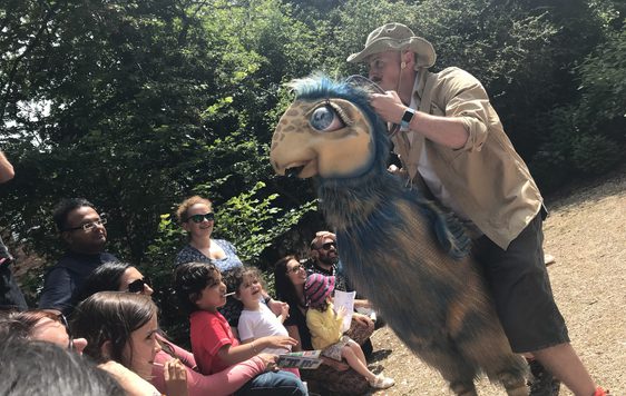 A person holding a cream and blue dinosaur puppet infront of a audience of people.