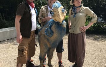 Three people dressed as explorers, one is holding a cream and blue dinosaur puppet.