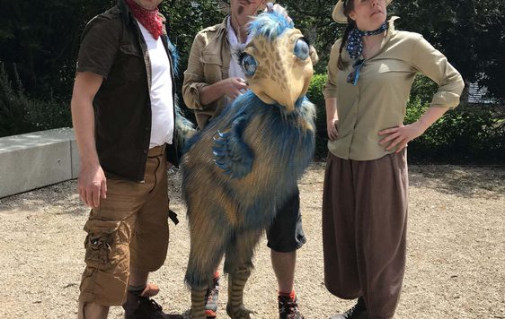 Three people dressed as explorers, one is holding a cream and blue dinosaur puppet.
