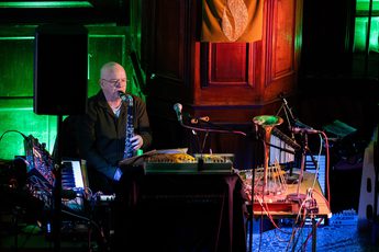 A person playing an instrument, and surrounded by keyboards in a dark room lit with coloured lights.