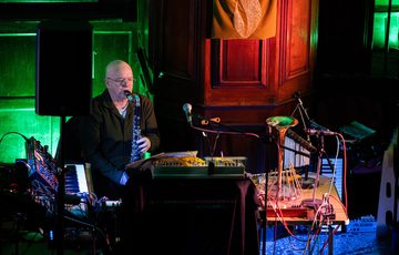 A person playing an instrument, and surrounded by keyboards in a dark room lit with coloured lights.