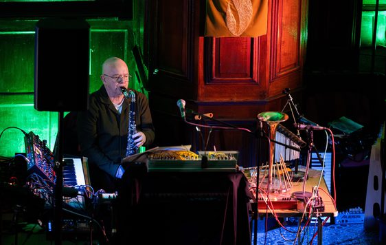 A person playing an instrument, and surrounded by keyboards in a dark room lit with coloured lights.