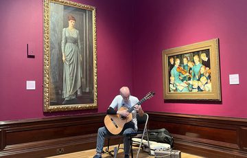 A person playing a guitar in a corner of a gallery with paintings in the background.