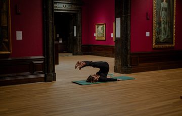 Person practicing pilates pose on mat inside an art gallery with framed paintings.