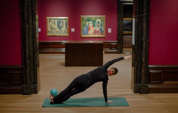 Person performing side plank pose on mat inside an art gallery with paintings.
