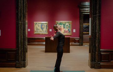 A woman standing performing a pilates pose with arms stretched out in an art gallery.