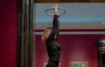 A person doing pilates holding a circular ring overhead against a gallery wall.
