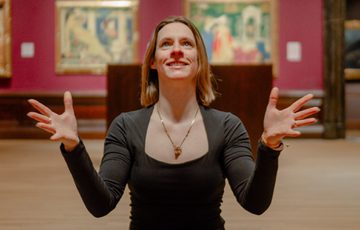 A woman sits cross-legged, smiling, tossing a green ball inside an art gallery room.
