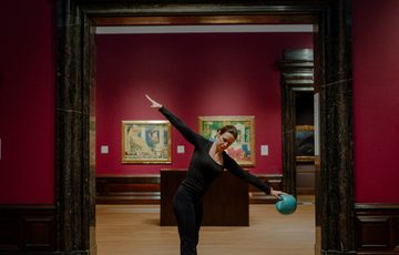 A woman balances on one leg, holding a turquoise ball, framed by a museum doorway.