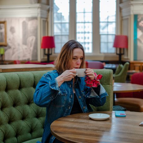 Woman in denim jacket drinks coffee at round wooden table in cozy café with green seating.
