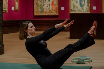A woman practices pilates on a mat in an art gallery with classical paintings in the background.