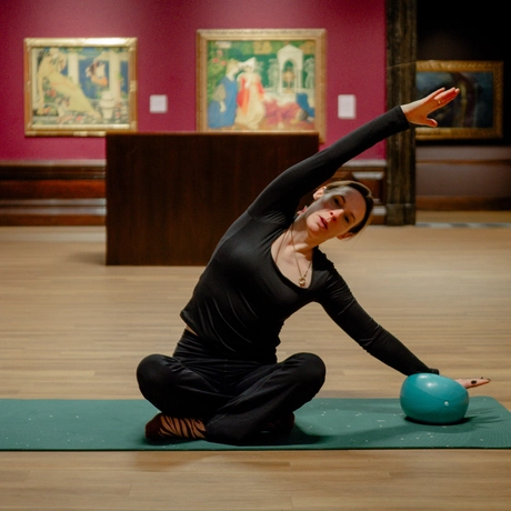 Woman practicing pilates on mat, stretching arm beside teal ball, inside art gallery.