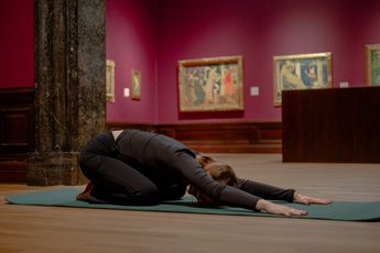 Person practicing yoga in child's pose on mat inside art gallery with framed paintings.