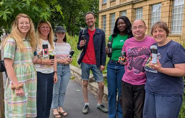 A group of smiling people holding camera's made out of cans, they are outside in front of a building.