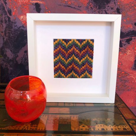 Close up of wool tapestry pattern on display in a white frame with red glass cup in front.