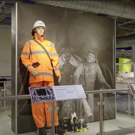 A display with mannequin wearing orange work clothes, with large black and white photo behind and label in front.