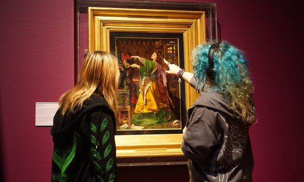 Two students look at a painting in a gold frame that is on display in an art gallery.
