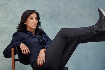 Picture of a lady against a grey background in a dark blue top and black trousers sitting with her feet raised against a wall
