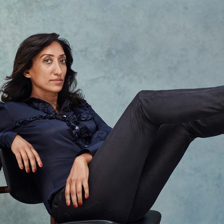 Picture of a lady against a grey background in a dark blue top and black trousers sitting with her feet raised against a wall