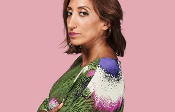 A picture of a lady wearing a green top with white and blue swirls, standing in front of a pink background