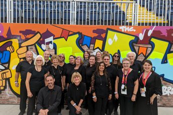 A photograph of a group of singers wearing black, standing in front of a wall in the background covered in street art