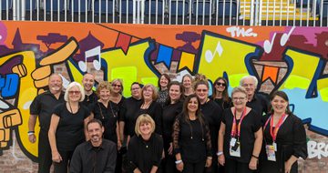 A photograph of a group of singers wearing black, standing in front of a wall in the background covered in street art