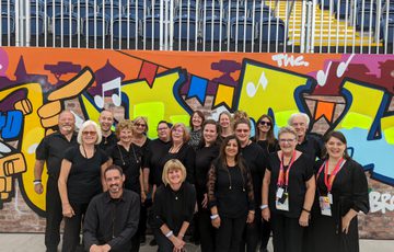 A photograph of a group of singers wearing black, standing in front of a wall in the background covered in street art
