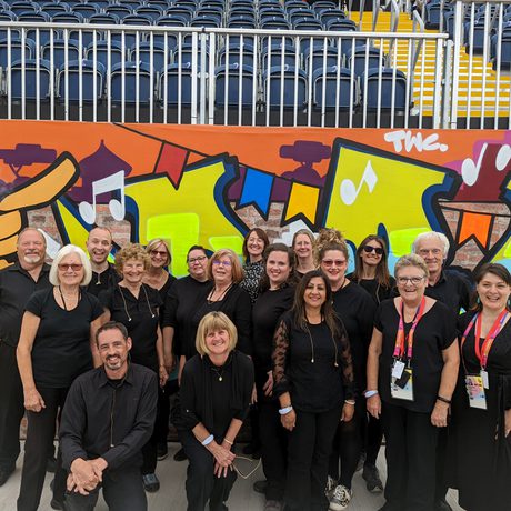A photograph of a group of singers wearing black, standing in front of a wall in the background covered in street art