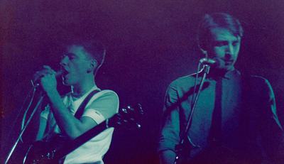 Two band members playing music, the vocalist is singing into the microphone, both hands gripping the mic. The other is looking downwards as he plays his guitar.