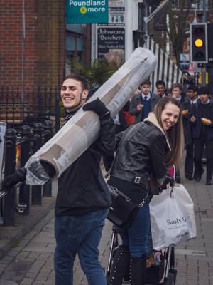 A man is carrying a rug over a shoulder. He is turned to look backwards, smiling. A woman next to him is pushing a pushcair, she is also looking backwards and smiling.