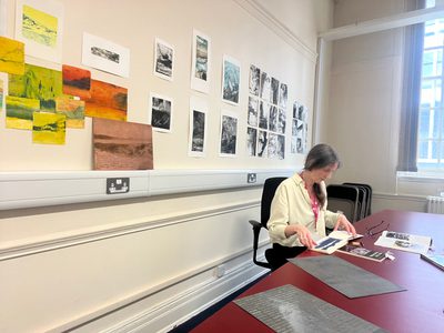 Fae in her studio space sitting at a desk with artworks on the wall behind.